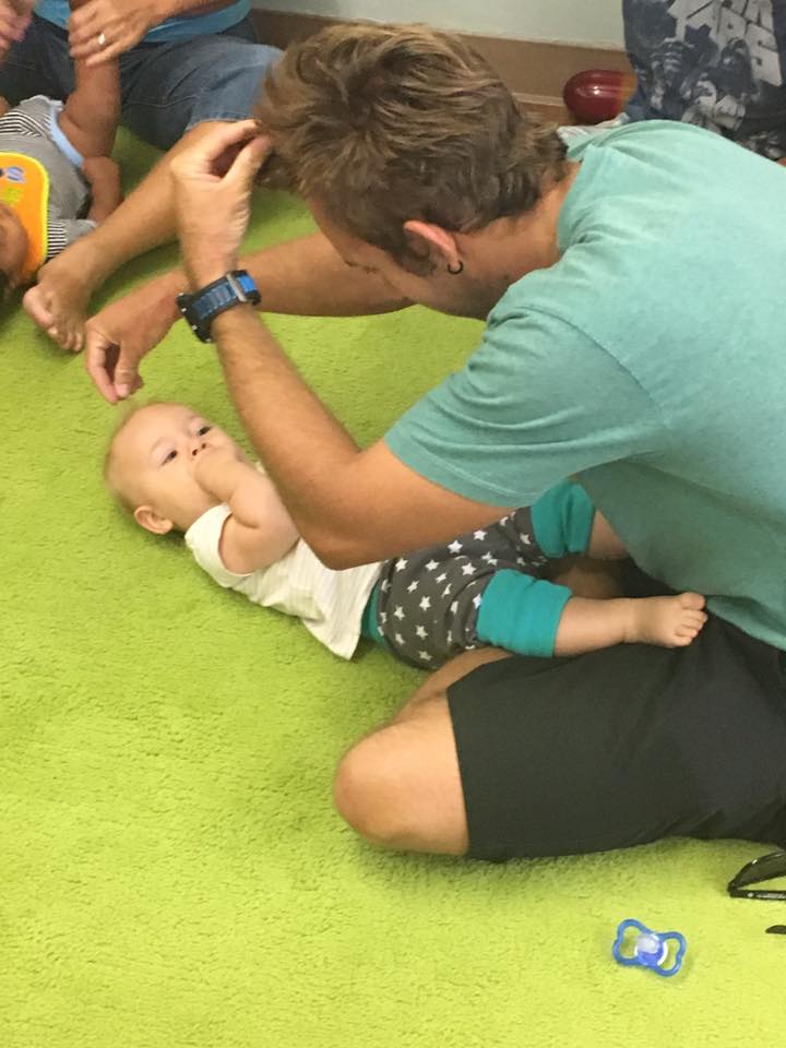 baby lying on a green carpet looking up at his father who is signing hair on himself and his son. Text: Baby Signs